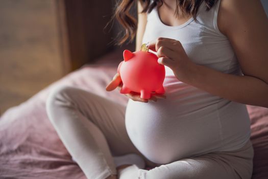 Pregnant woman relaxing at home. She is sitting on bed and saving coin in piggy bank.