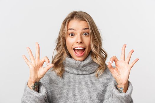 Close-up of attractive blond girl, showing okay signs and smiling excited, standing over white background.