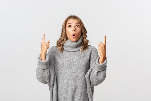 Image of surprised and happy blond girl, wearing grey sweater, pointing fingers up and looking impressed, white background.