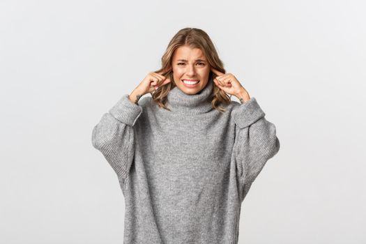 Portrait of annoyed blond girl in grey sweater complaining on loud noise, shut her ears with fingers and grimacing bothered, standing over white background.