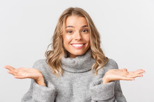 Close-up of cute blond girl in grey sweater, shrugging and smiling, dont know anything, standing over white background.