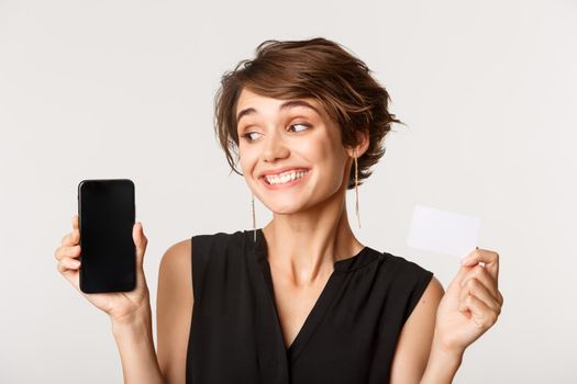 Close-up of beautiful cheerful young woman showing credit card, looking at mobile phone screen, standing over white background.