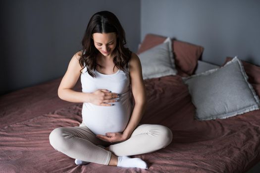Happy pregnant woman relaxing at home. She is sitting on bed in bedroom.