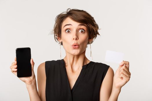 Close-up of amazed young woman showing credit card and smartphone screen, looking with interest, standing over white background.