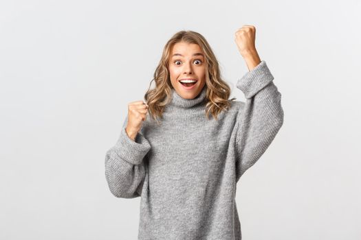 Image of excited hopeful girl in grey sweater, looking surprised as winning and rejoicing, raising hand up, standing happy against white background.