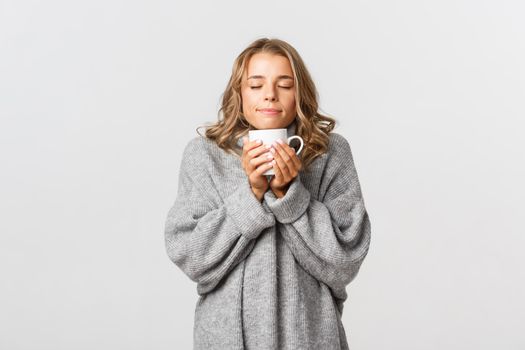 Portrait of beautiful blond girl in grey sweater, smelling coffee from cup, standing over white background.