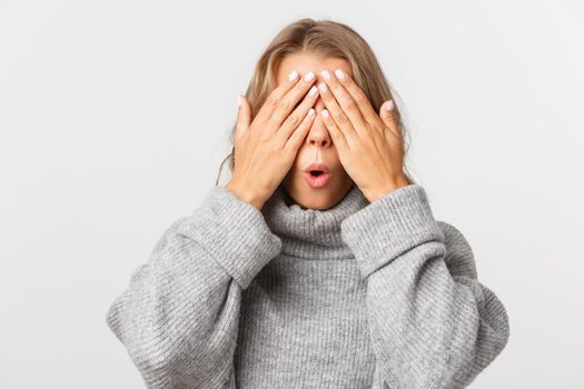 Close-up of happy beautiful woman in grey sweater, standing blindsided with hands on face, waiting for surprise, white background.