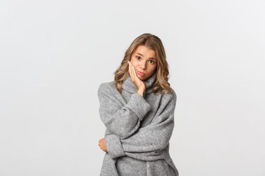 Portrait of sad beautiful female model in grey sweater, looking at camera with gloomy face, standing over white background.