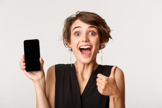 Cheerful beautiful girl showing thumbs-up and demonstrating smartphone screen, standing over white background.