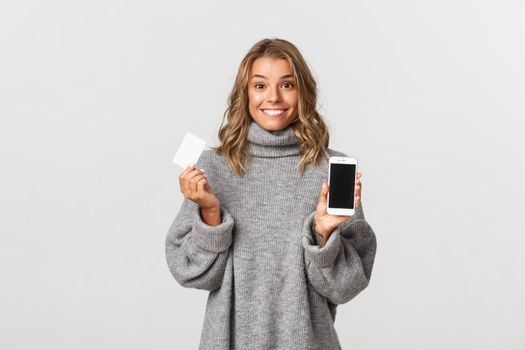 Pretty blond girl in grey sweater, showing smartphone screen with credit card for shopping online, standing against white background.