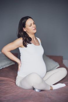 Pregnant woman is having pain in her back. She is sitting on bed at home.