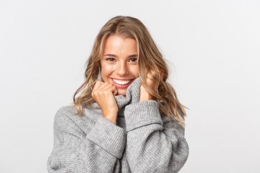 Close-up of attractive flirty woman in grey sweater, smiling confident at camera, standing over white background.