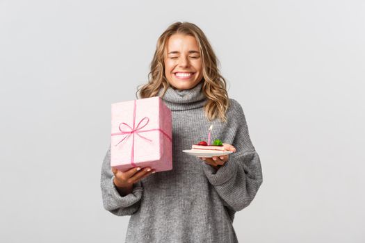 Image of happy attractive girl making wish and celebrating birthday, holding b-day cake and gift, standing against white background.