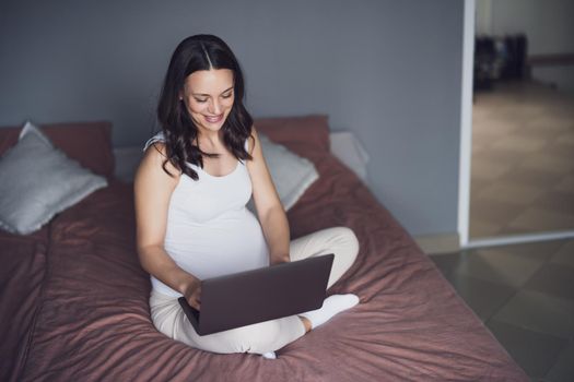 Happy pregnant woman relaxing at home. She is sitting on bed in bedroom and using laptop.