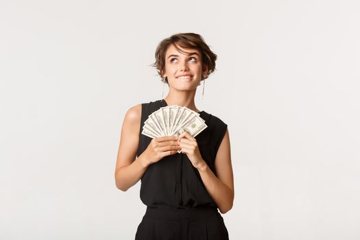 Dreamy beautiful girl looking upper left corner and thinking, holding money, standing over white background.