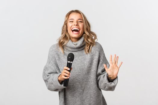 Studio shot of happy blond girl in grey sweater, singing karaoke in microphone and smiling, standing over white background.