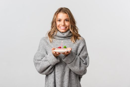 Lovely birthday girl in grey sweater smiling, holding b-day cake and making wish, standing over white background.