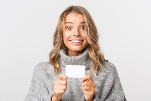 Close-up of excited smiling woman with blond hair, holding credit card for shopping, standing over white background.