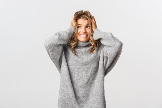 Portrait of blond girl in panic, looking left and holding hands on head, feeling alarmed and distressed, standing over white background.