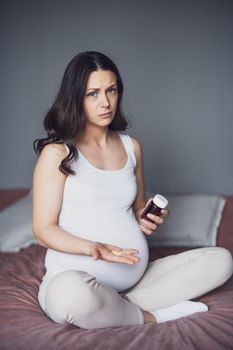 Pregnant woman is sitting on bed and holding pills. She is unwell.