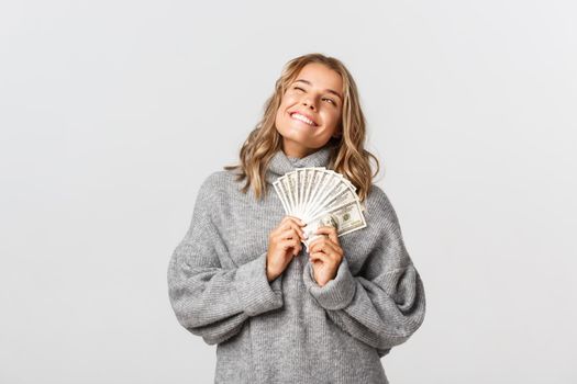 Cheerful blond girl in grey sweater, smiling pleased, holding money, standing over white background.