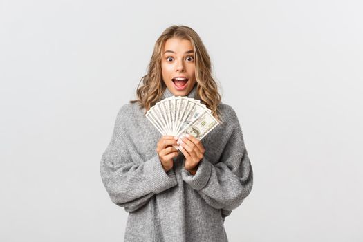 Image of excited attractive girl in grey sweater, holding money and looking amazed, standing over white background.