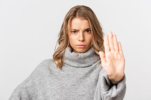 Close-up of serious and confident blond girl in grey sweater, frowning and showing stop gesture, prohibit something, disapprove and forbid action, white background.