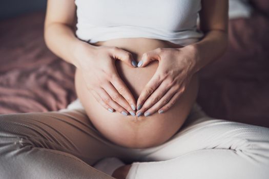 Pregnant woman relaxing at home. She is making heart shape on her stomach.