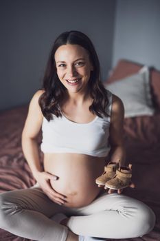 Pregnant woman relaxing at home. She is sitting on bed and holding baby shoes.