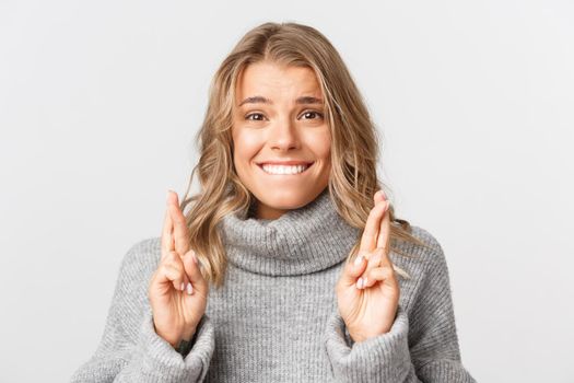Close-up of beautiful hopeful girl with short blond hairstyle, wearing grey sweater, crossing fingers and making wish, standing over white background.