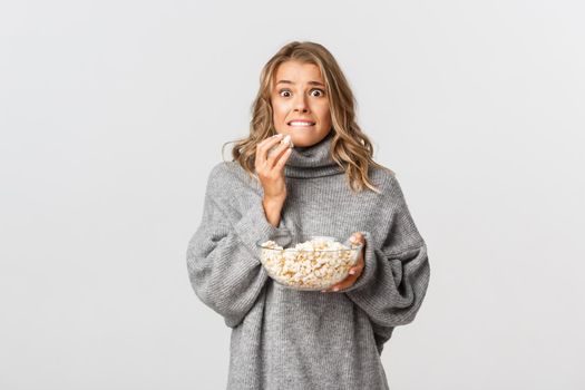 Portrait of blond girl eating popcorn and looking scared, watching movie, standing over white background.