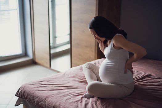 Pregnant woman is having pain in her stomach and back. She is sitting on bed at home.