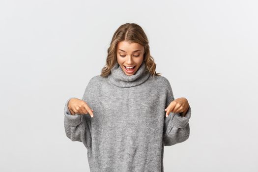 Cheerful blond girl rejoicing as looking and pointing fingers down at your logo banner, standing in grey sweater over white background.