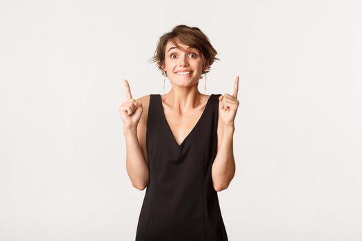 Hopeful girl pointing fingers up and smiling, looking at camera excited, standing over white background.