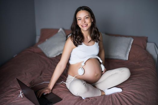 Happy pregnant woman relaxing at home. She is sitting on bed in bedroom and playing music to her baby in stomach.