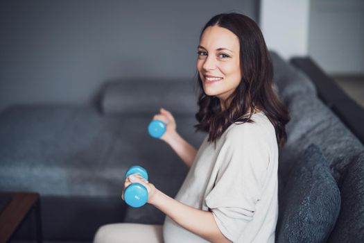 Portrait of pregnant happy woman who is exercising with weights at home.