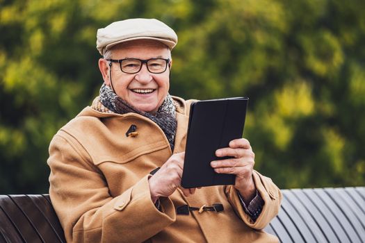 Outdoor portrait of cheerful senior man in winter coat who is using digital tablet.
