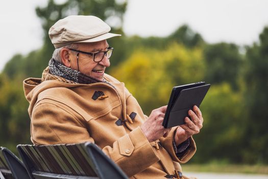 Outdoor portrait of cheerful senior man in winter coat who is using digital tablet.