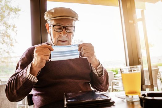 Senior man is sitting in cafe with face protection mask. Covid 19 prevention concept.