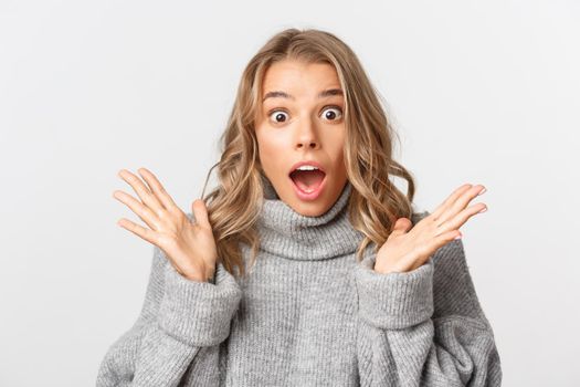 Close-up of shocked gasping girl in grey sweater, open mouth amazed, standing over white background.