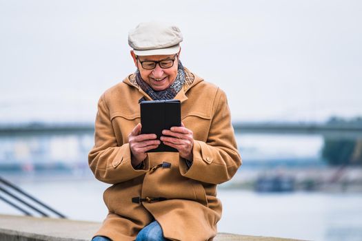 Outdoor portrait of cheerful senior man in winter coat who is using digital tablet.