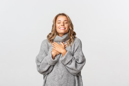 Close-up of beautiful blond girl, wearing grey sweater, feeling nostalgic, holding hands on heart and close eyes, daydreaming over white background.