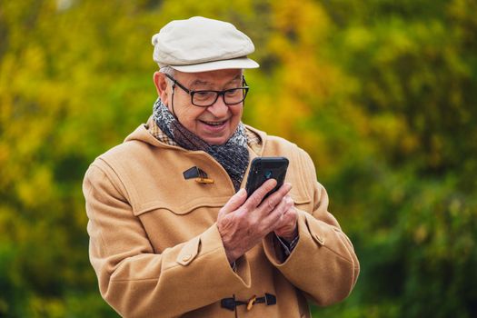 Outdoor portrait of cheerful senior man in winter coat who is using smartphone.