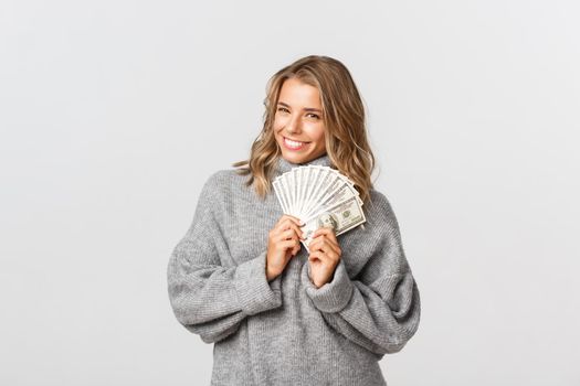 Image of beautiful blond girl in grey sweater, holding money and smiling, thinking how to waste cash, white background.