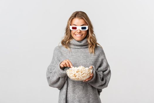 Young pretty girl with blond short hairstyle, wearing 3d glasses, watching movie and eating popcorn, standing over white background.