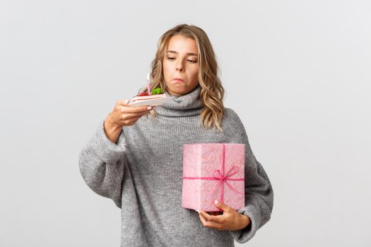 Studio shot of cute blond girl holding gift, looking curious at birthday cake with lit candle, standing over white background.