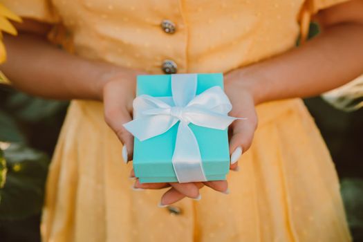 Woman holding blue gift box with bow in sunflowers field. Present, bonus, offer concept. Only hands. Warm yellow toning. Trendy . High quality photo