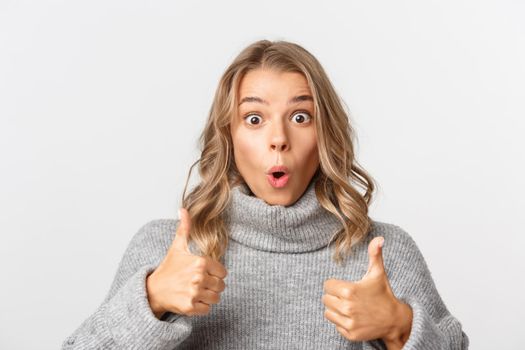 Close-up of impressed cute girl in grey sweater, showing thumbs-up and like something, praising good choice, standing over white background.