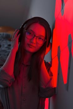 Young smiling woman with wireless headphones in red light of neon panels leans on wall listening to music, nightclub concept, futuristic neon trend of modern electronic sound, dark vertical image