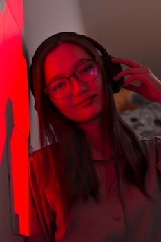 Young smiling pretty woman with wireless headphones in red light of neon panels leans on wall looking at lens, nightclub concept, futuristic neon trend of modern electronic music, dark vertical image
